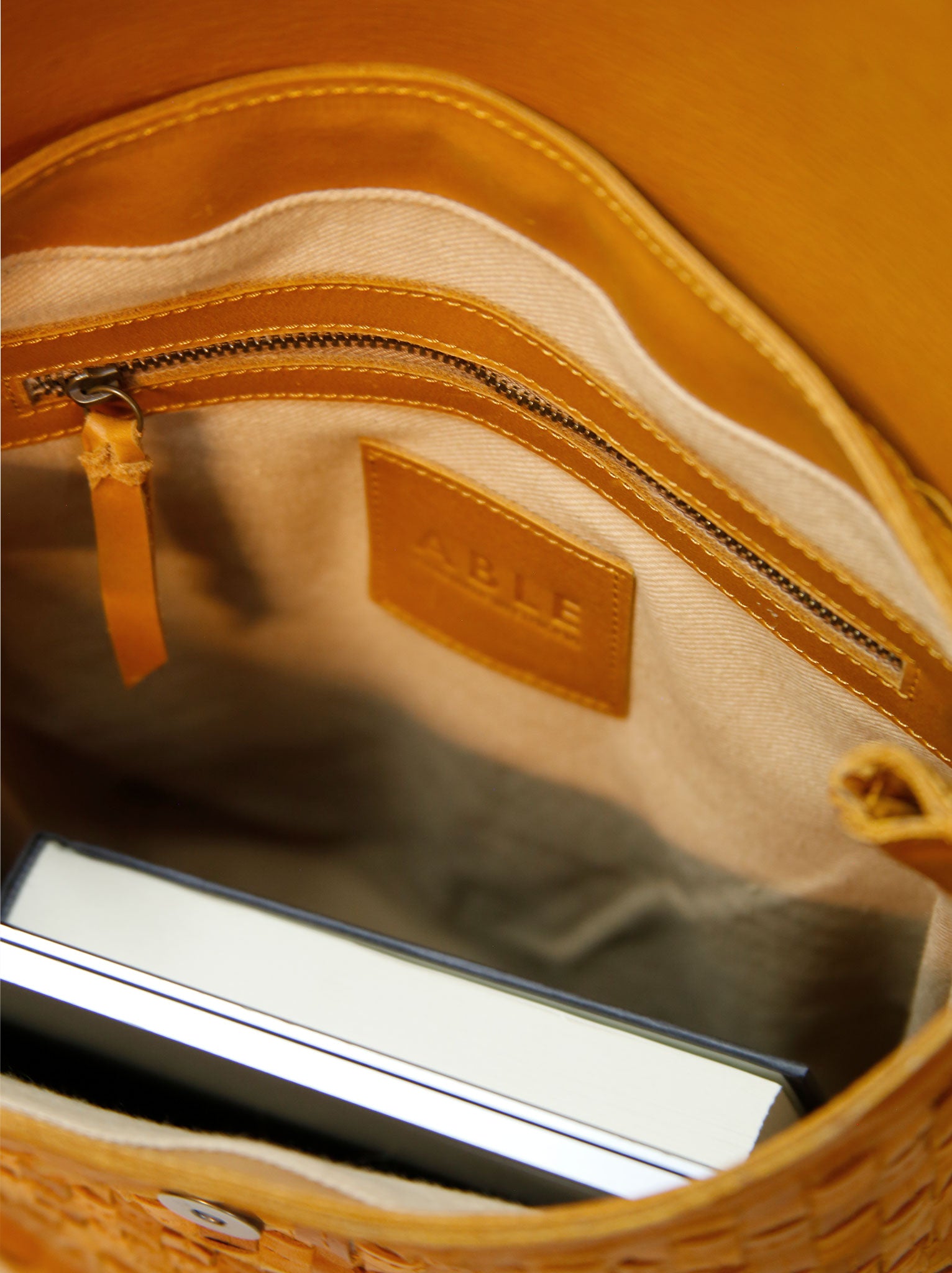 Tan lined interior of basketweave backpack with interior zipper pocket, with a book and laptop inside. #Color_Cognac