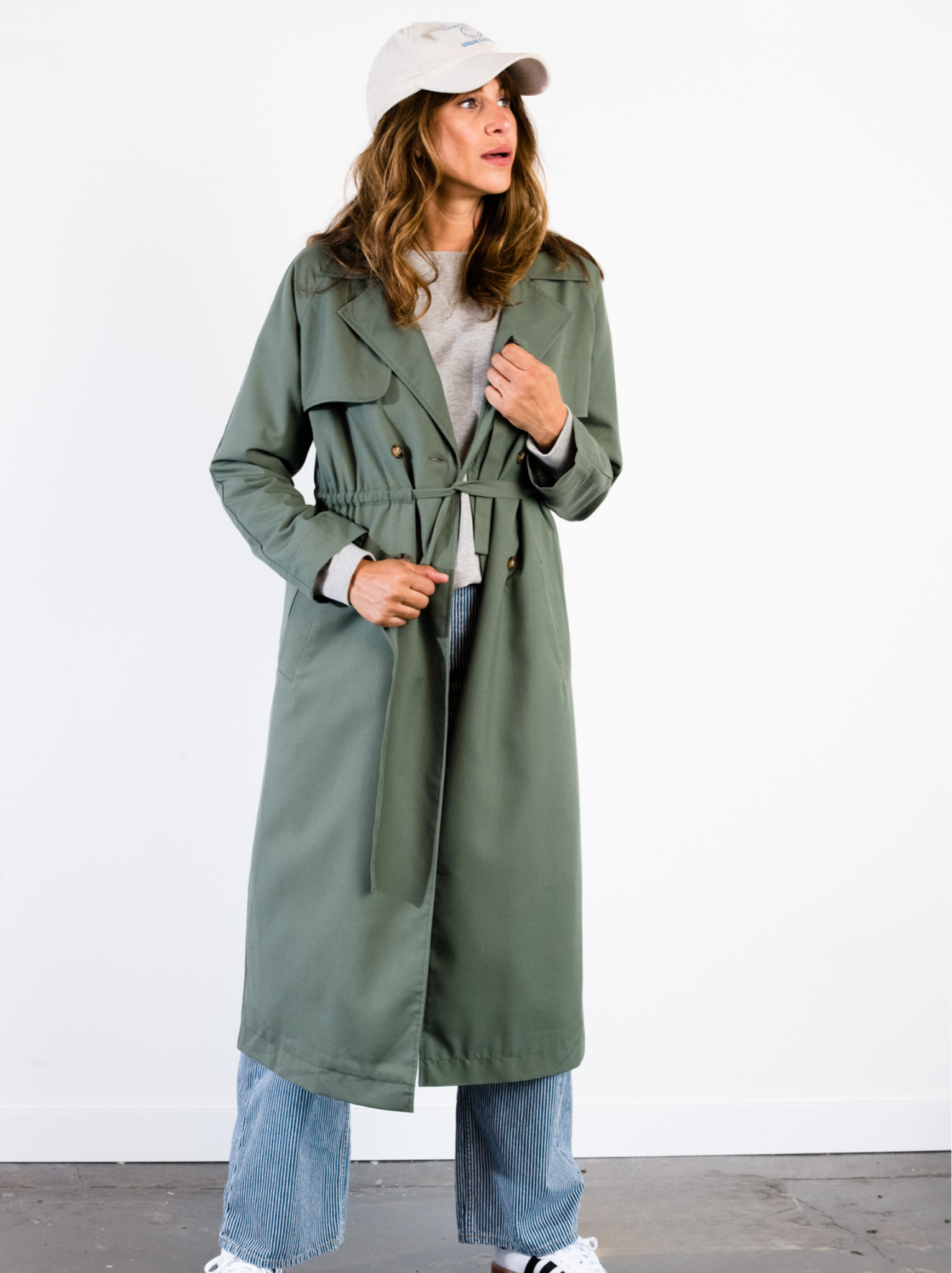 A woman tying the belt of a green long sleeve trench coat inside against neutral background. #Color_Moss