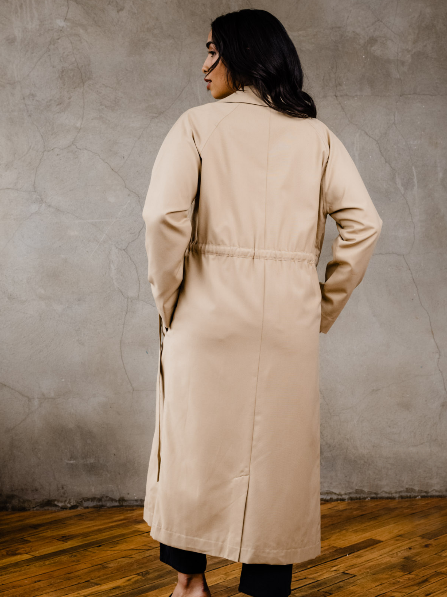 Back of woman wearing tan trench coat with hands in both pockets, indoors on wooden floor with a neutral background. #Color_Brown Sugar