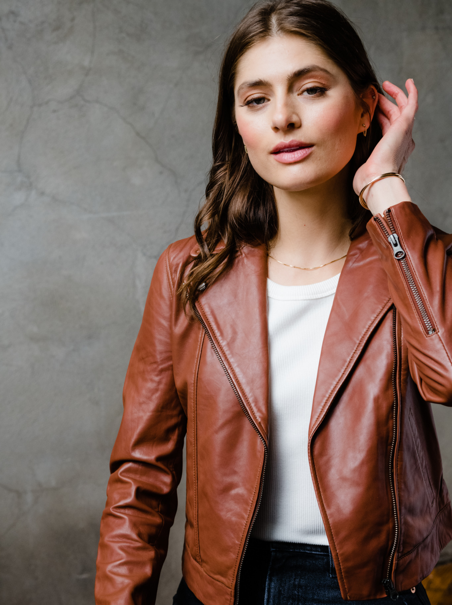 A woman wearing a brown leather jacket, open, with her arm up showing the sleeve zipper detail. #Color_Whiskey