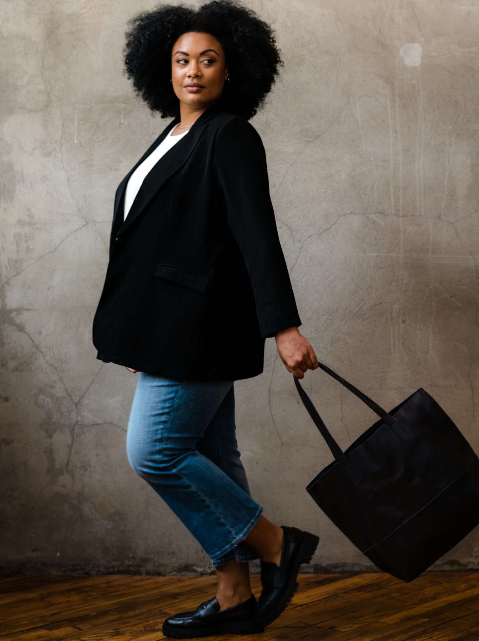 Woman holding the straps of a black leather tote in her hands as she walks on a wooden floor with a neutral background. #Color_Black