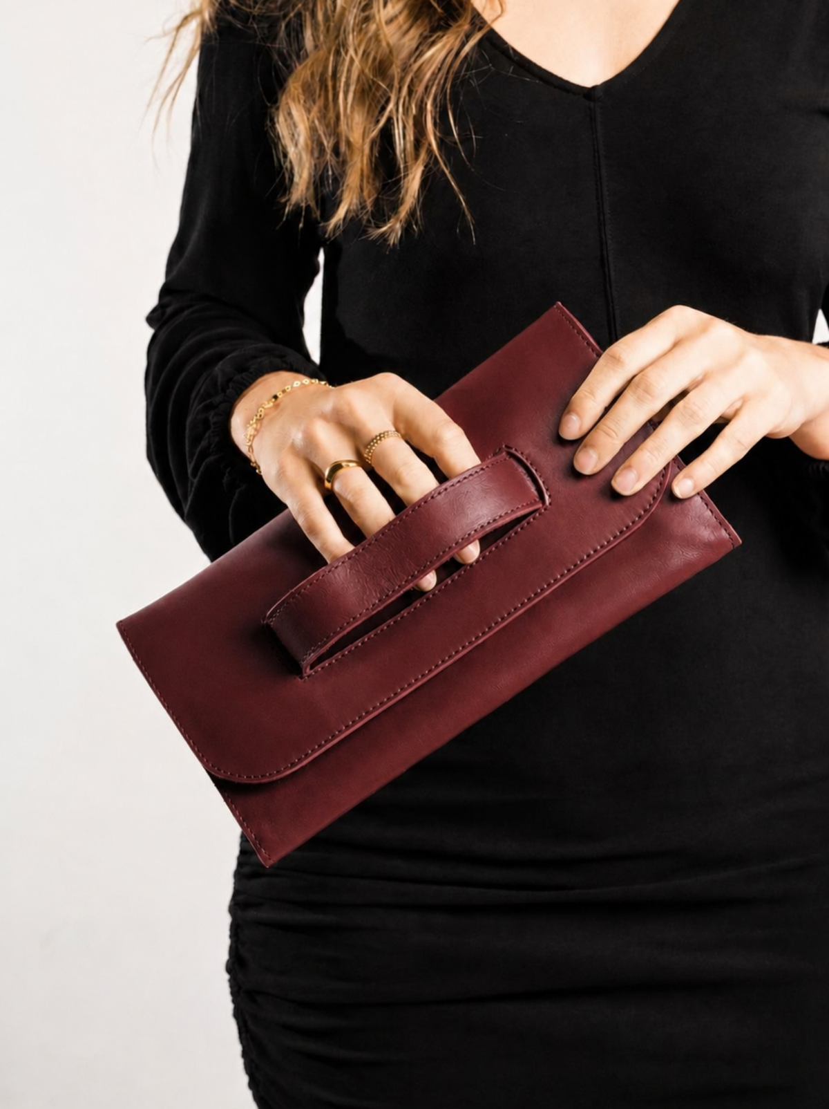 A person in a black long-sleeve dress holds a burgundy leather clutch bag with a front handle against a plain light background. #Color_Wine