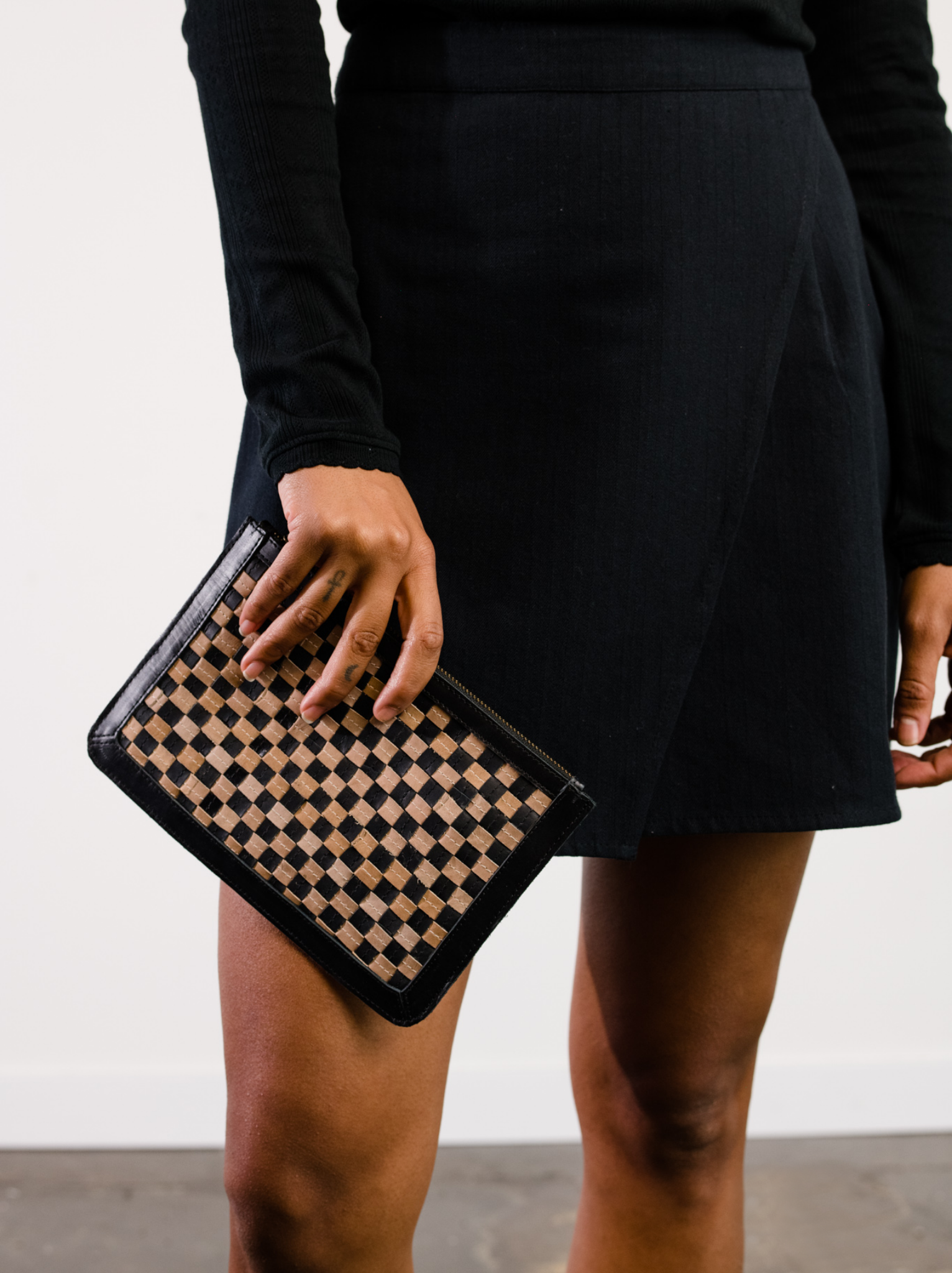 Woman holding  a black and tan checkered clutch, wearing a black skirt and top against a white wall. #Color_Black/Tan