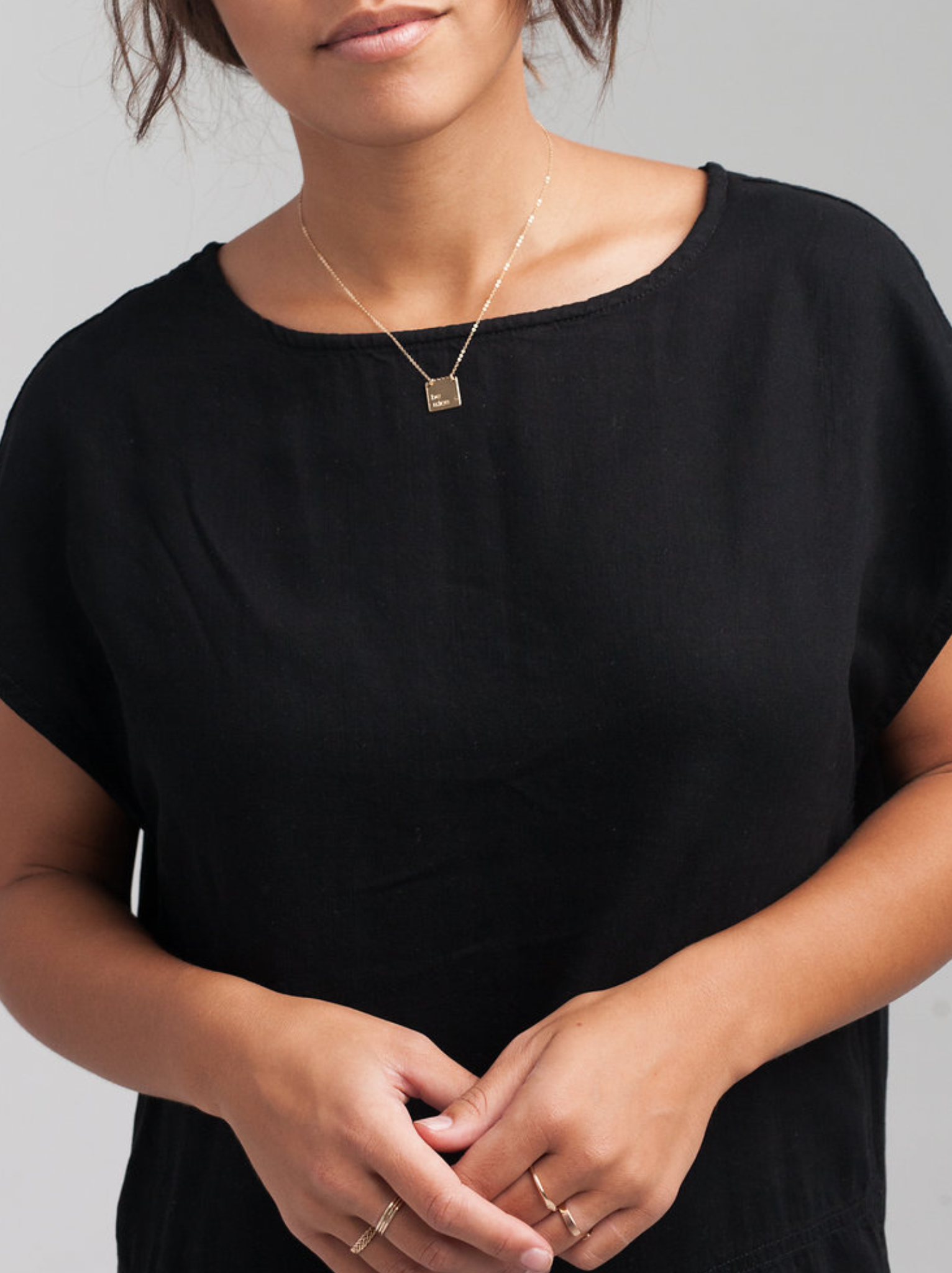 A woman wearing a gold necklace with a personalized square pendant against a white background.