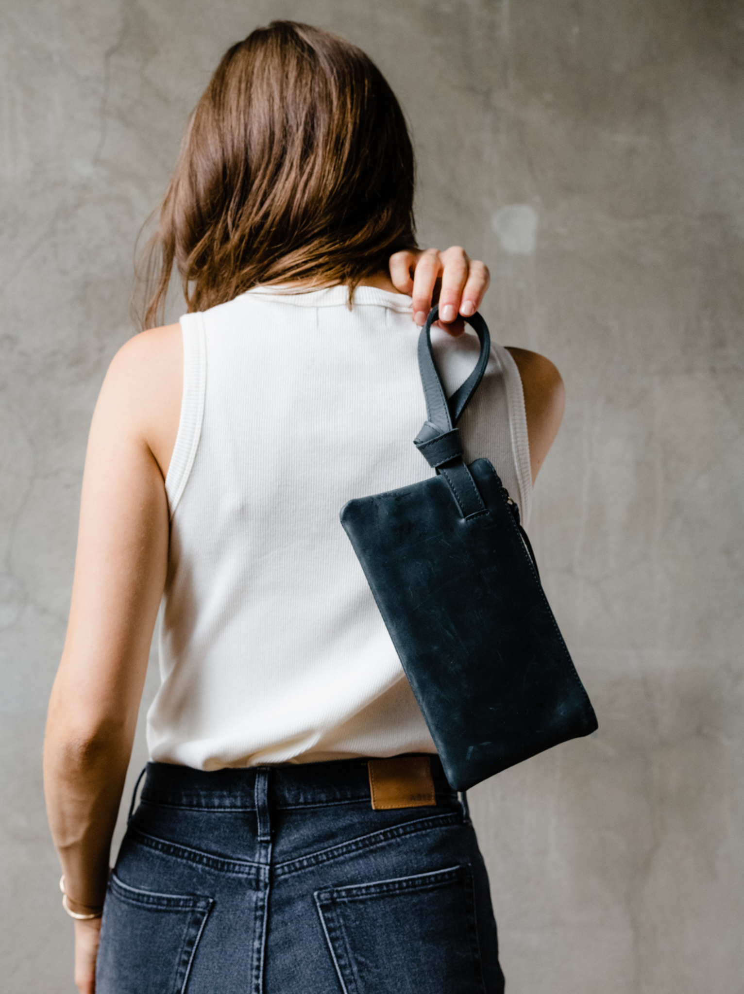 Woman holding a black leather wristlet by the handle over her back against neutral background. #Color_Black