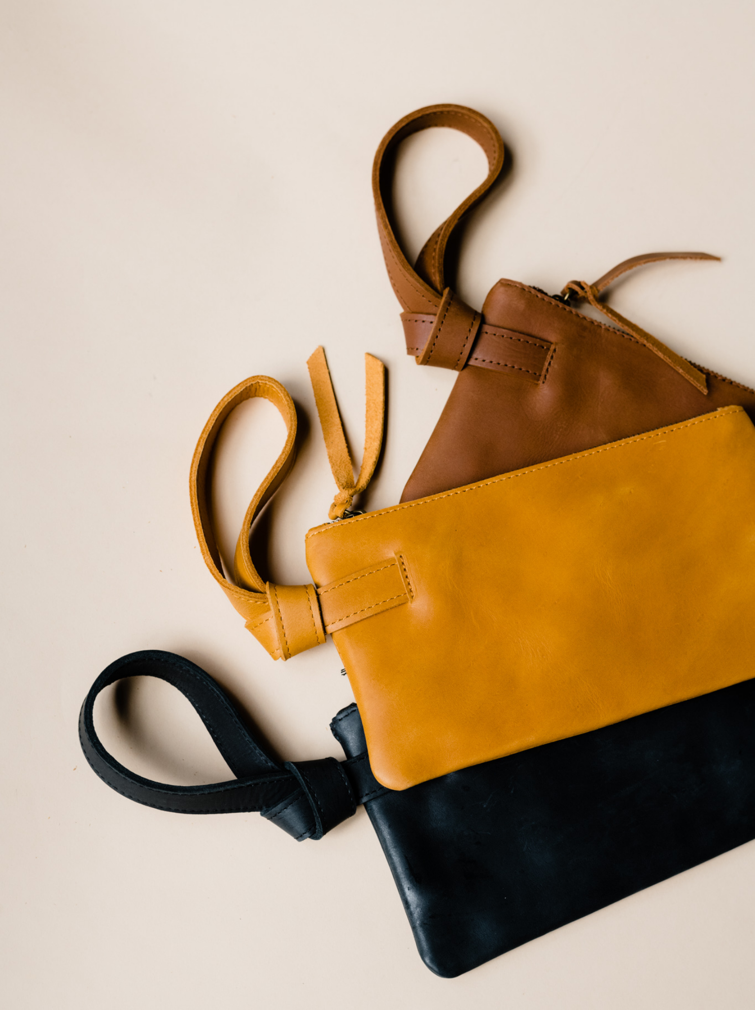 Three stacked leather wristlets, brown, orange, and black, against a tan background. #Color_Cognac