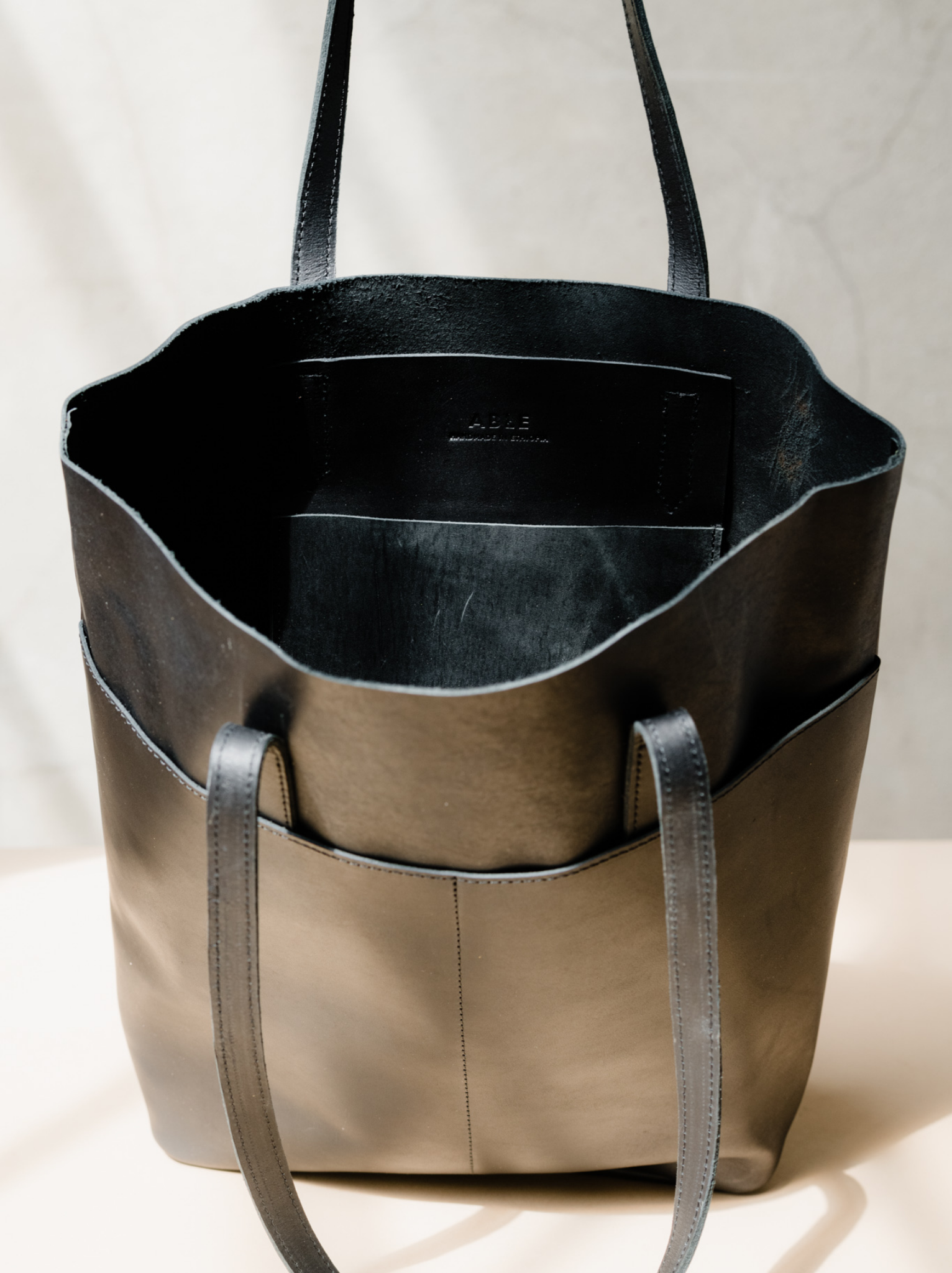 Unlined interior with black leather slip pocket of black tote on tan surface with neutral background. #Color_Black