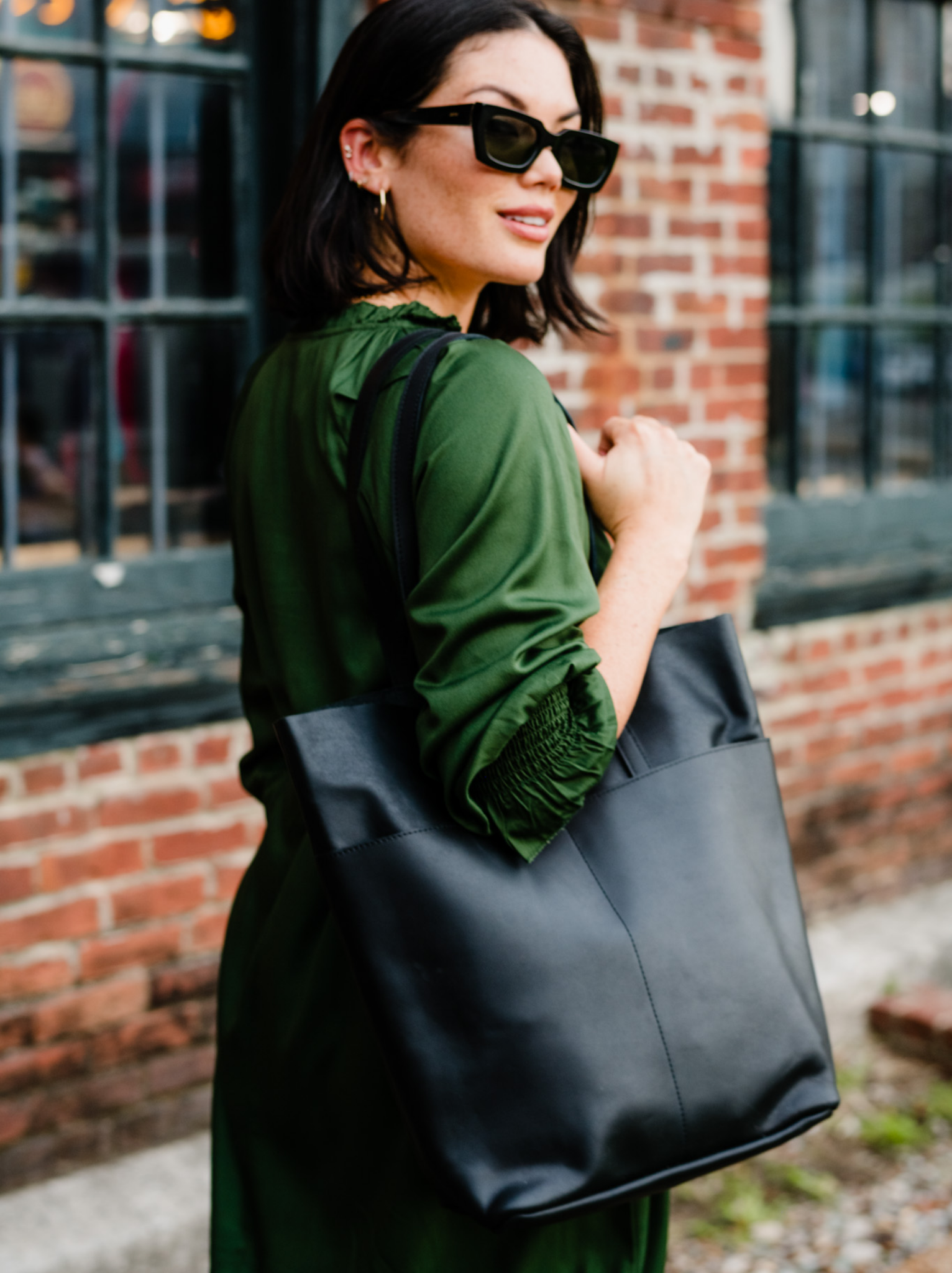 Woman outside holding black leather tote on her shoulder wearing a green dress and sunglasses against a brick wall with windows. #Color_Black