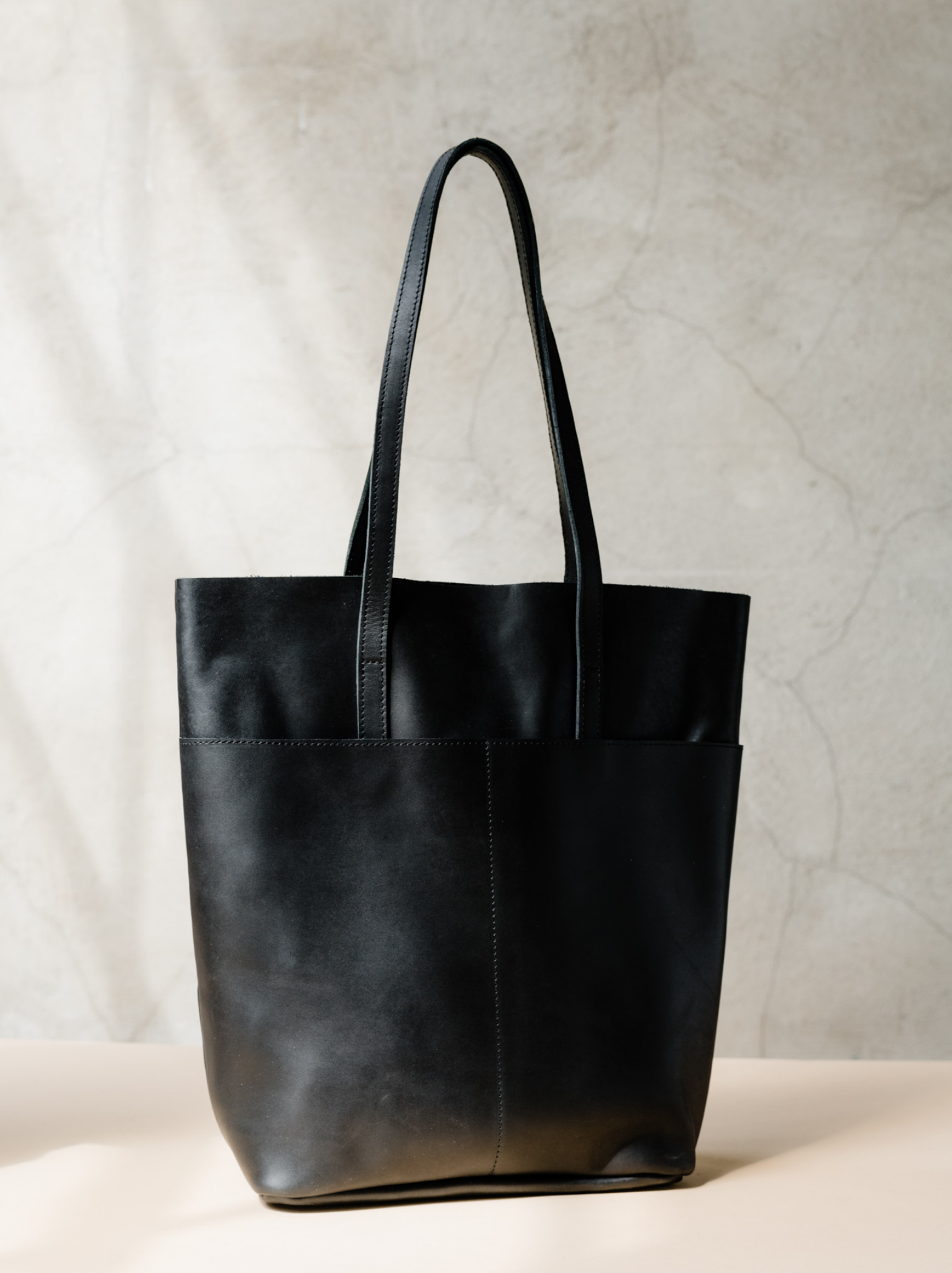 Black leather tote with shoulder straps and two slip pockets on a tan surface against a neutral background. #Color_Black
