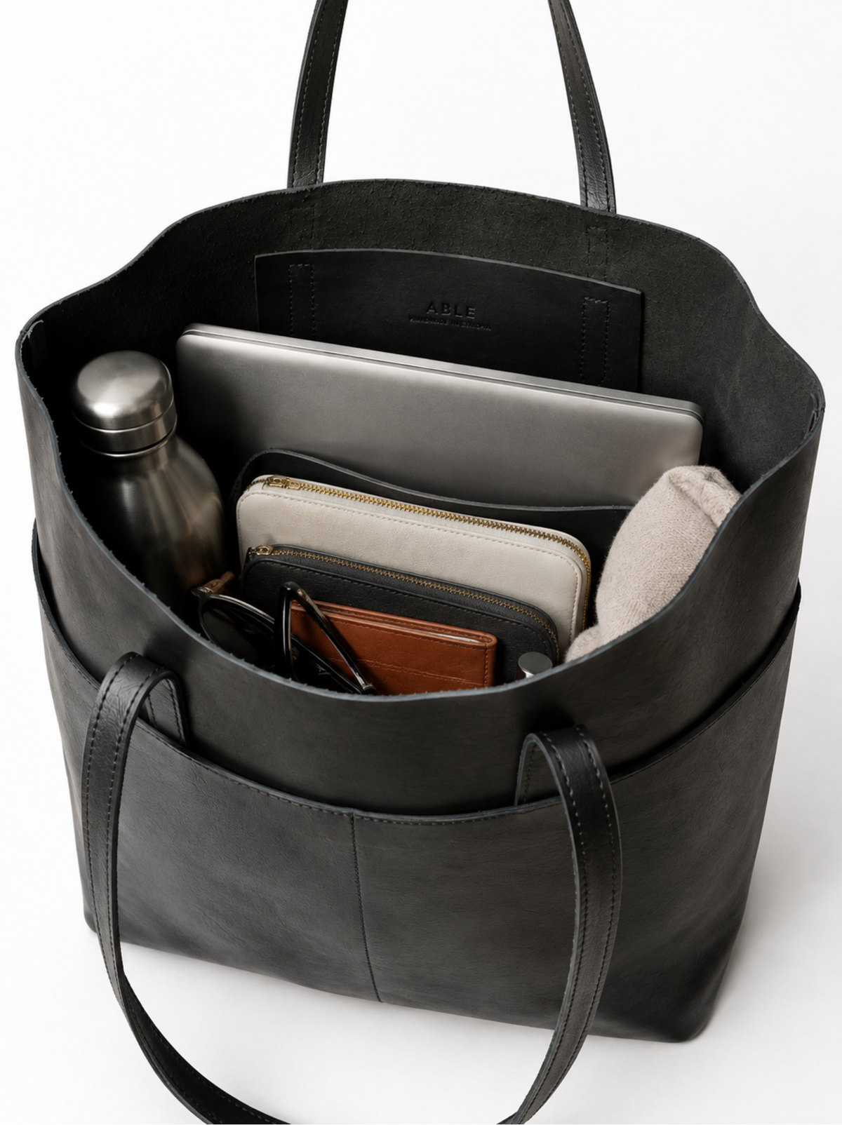 A black leather tote bag shown open from above, filled with travel accessories including a silver laptop, wallet, sunglasses, and a metal water bottle. The brand name 'ABLE' is embossed on the inner pocket. #Color_Black