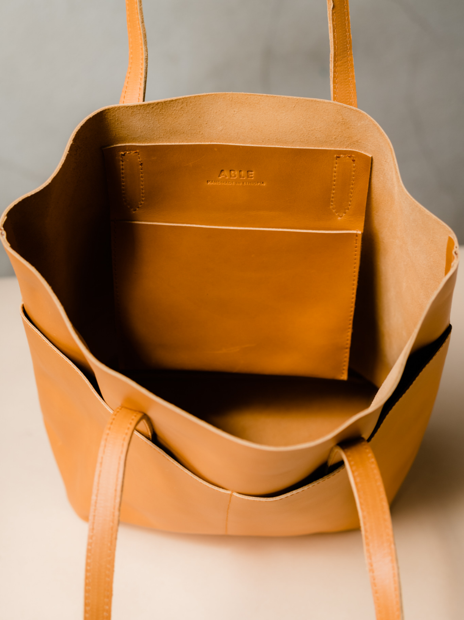 Unlined interior of an orange cognac leather tote bag with an interior slip pocket on a tan surface against a neutral background. #Color_Cognac