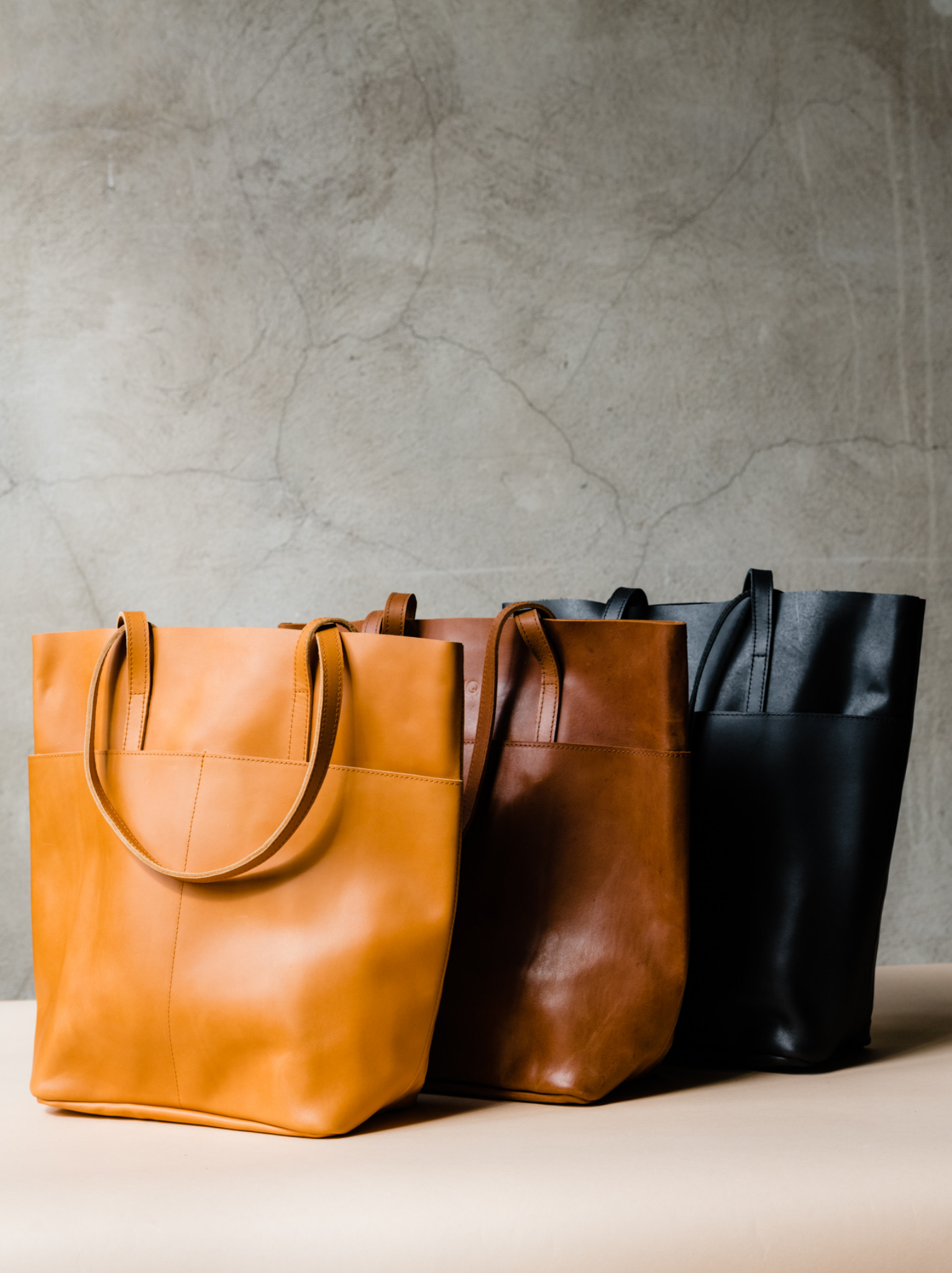 Three leather totes, orange cognac, brown, and black, lined up on a tan surface against a neutral background. #Color_Cognac