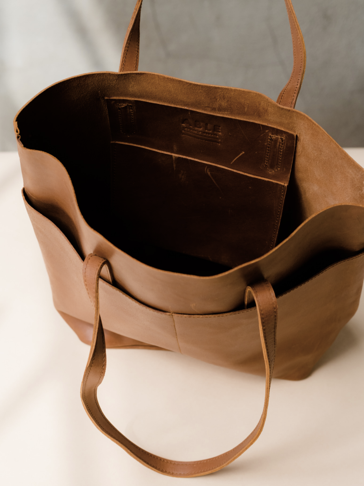 Unlined interior of a brown leather tote bag with an interior leather slip pocket on a tan surface against a neutral background. #Color_Whiskey