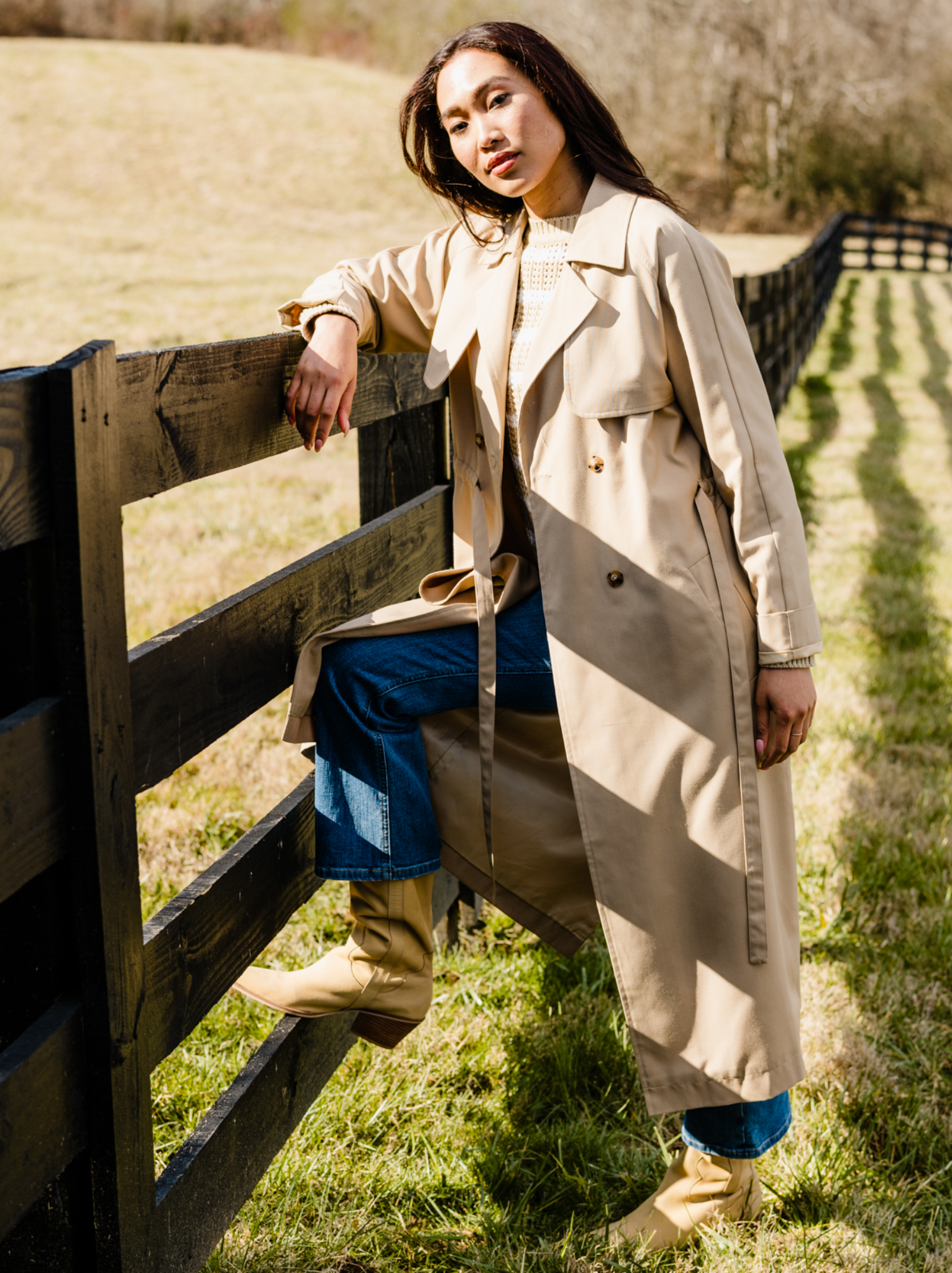 A woman standing next to a wooden fence, wearing a beige trench coat with long sleeves and pockets. #Color_Brown Sugar