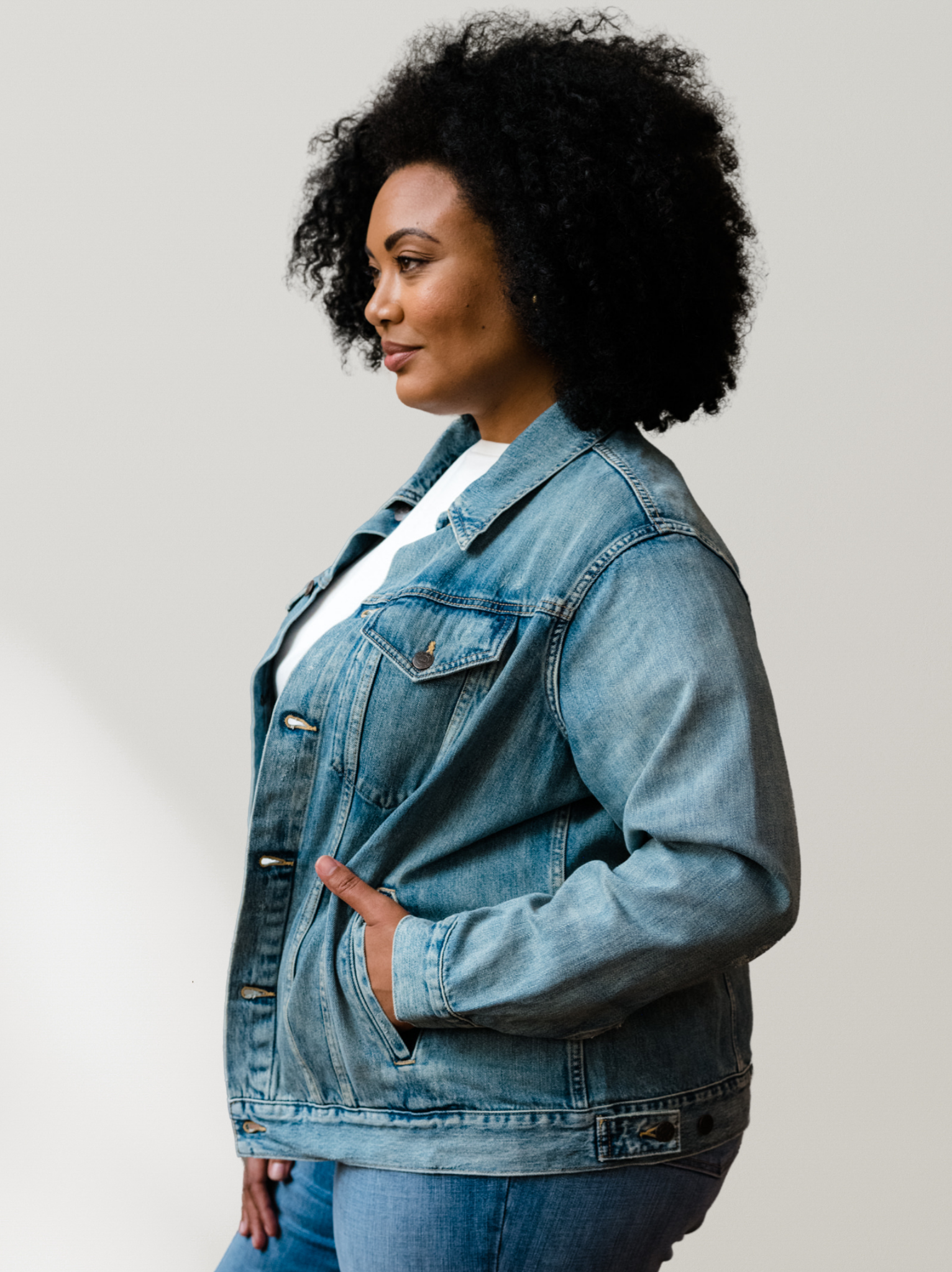 A side profile of woman wearing a light blue denim jacket with her hand partially in the jacket pocket against a neutral background. #Color_Original Wash