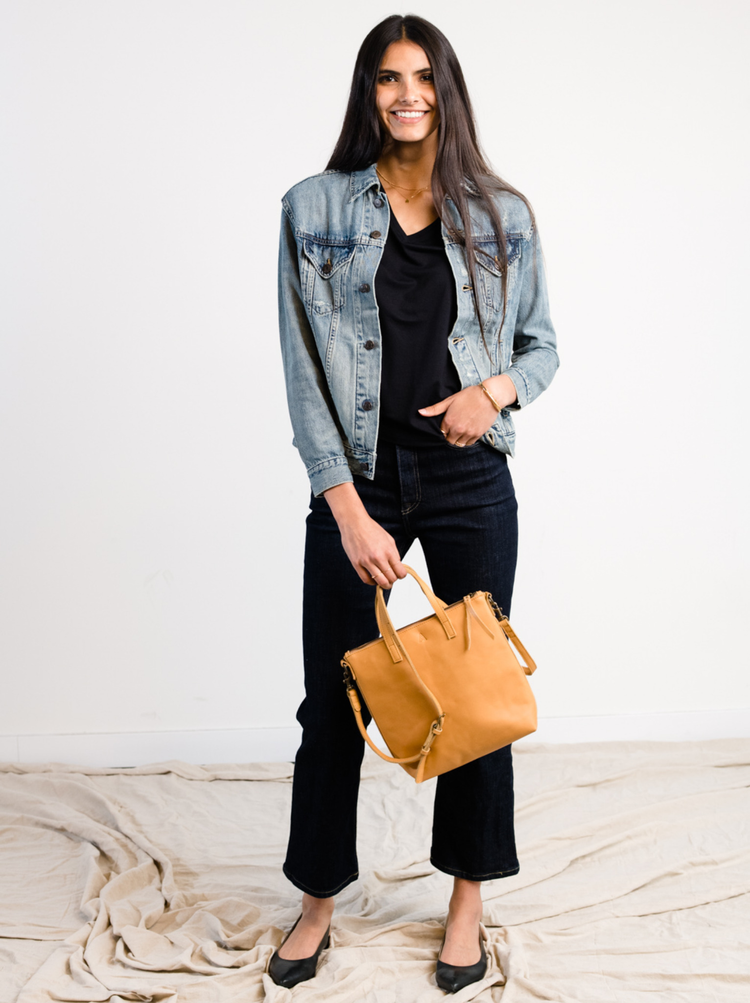 A woman wearing a light blue denim jacket with light distressing and holding a brown leather bag against a white background. #Color_Original Wash