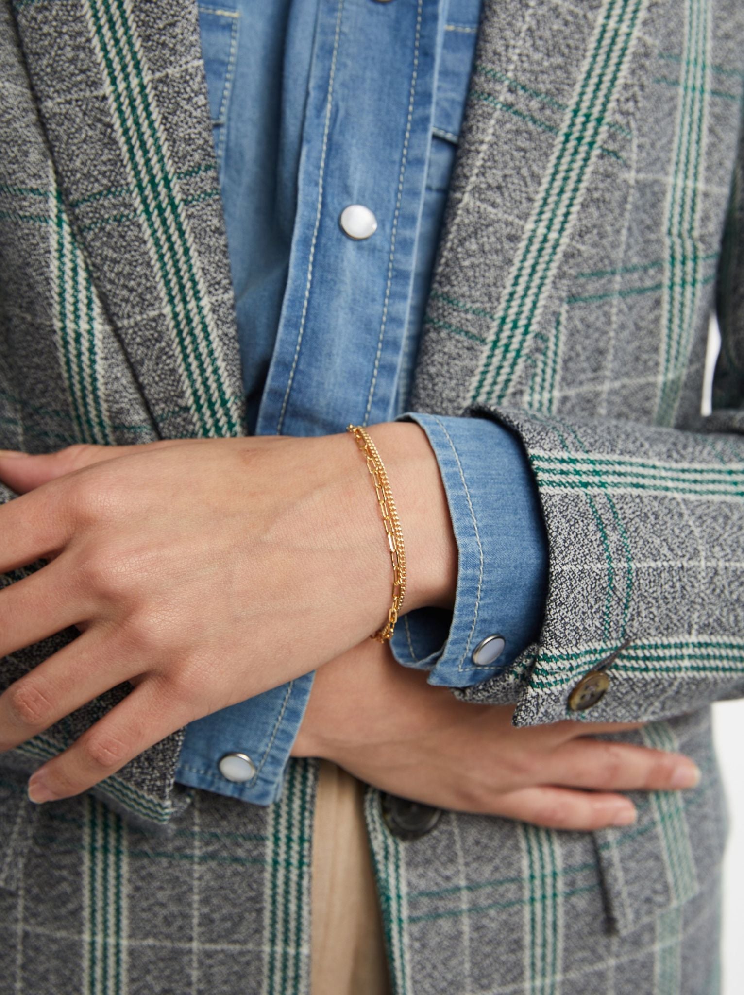 A close up of a woman wearing a gold layered chain bracelet on her wrist in a denim shirt and blazer.