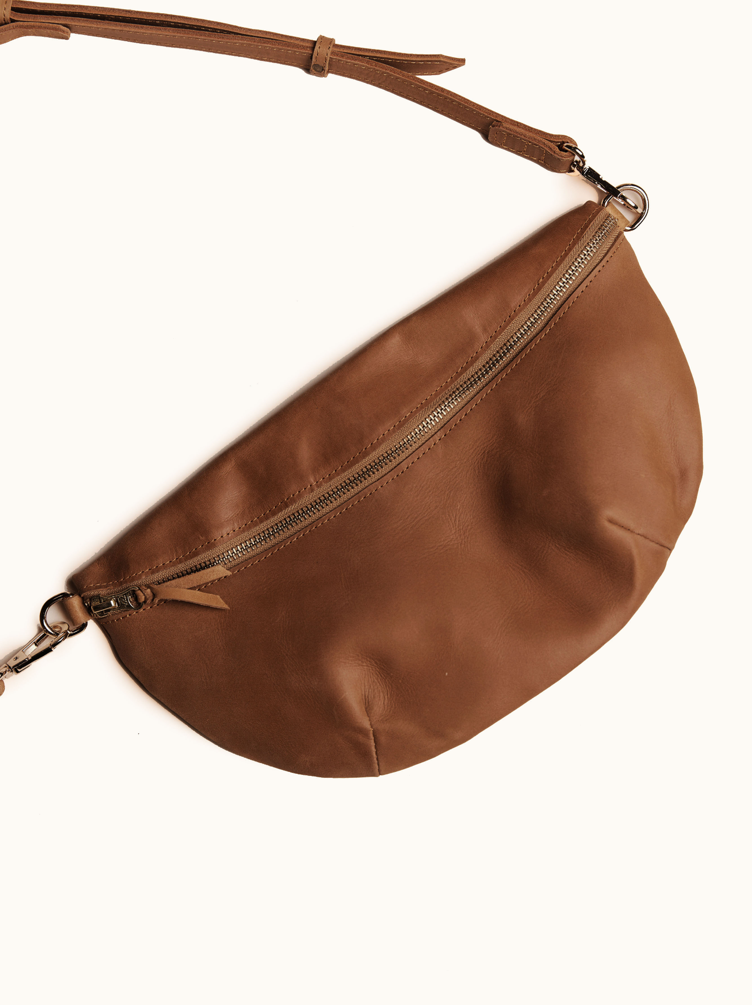 Brown leather zip up belt bag with metal hardware lying on a white background. #Color_Whiskey